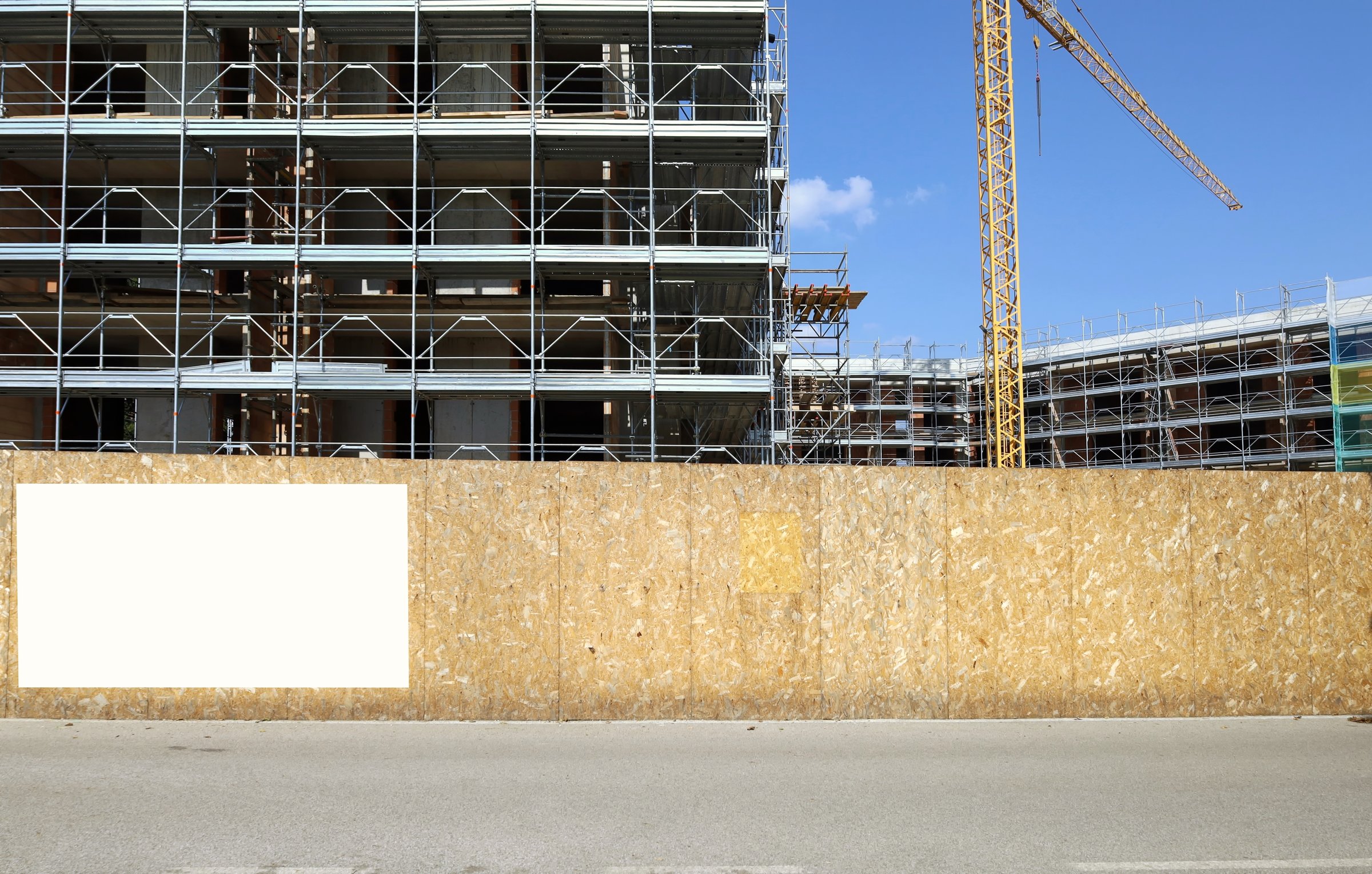Blank information board on fence outside the construction site with a new large building among the scaffolding, Tower crane and sky. Background for copy space.