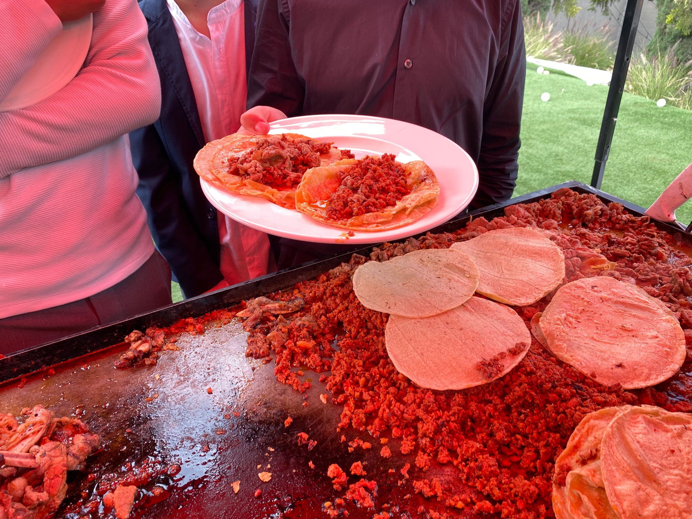 People serving tacos filled with seasoned meat from a griddle at an outdoor gathering.