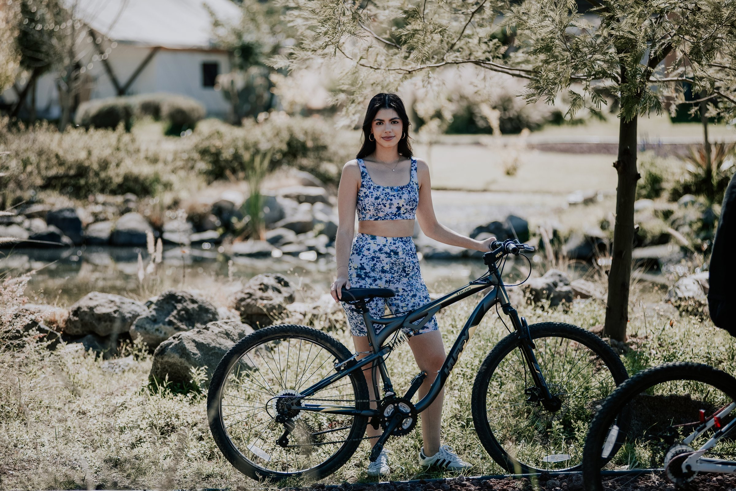 Woman in a blue floral outfit standing with a bicycle outdoors in a grassy area near trees.