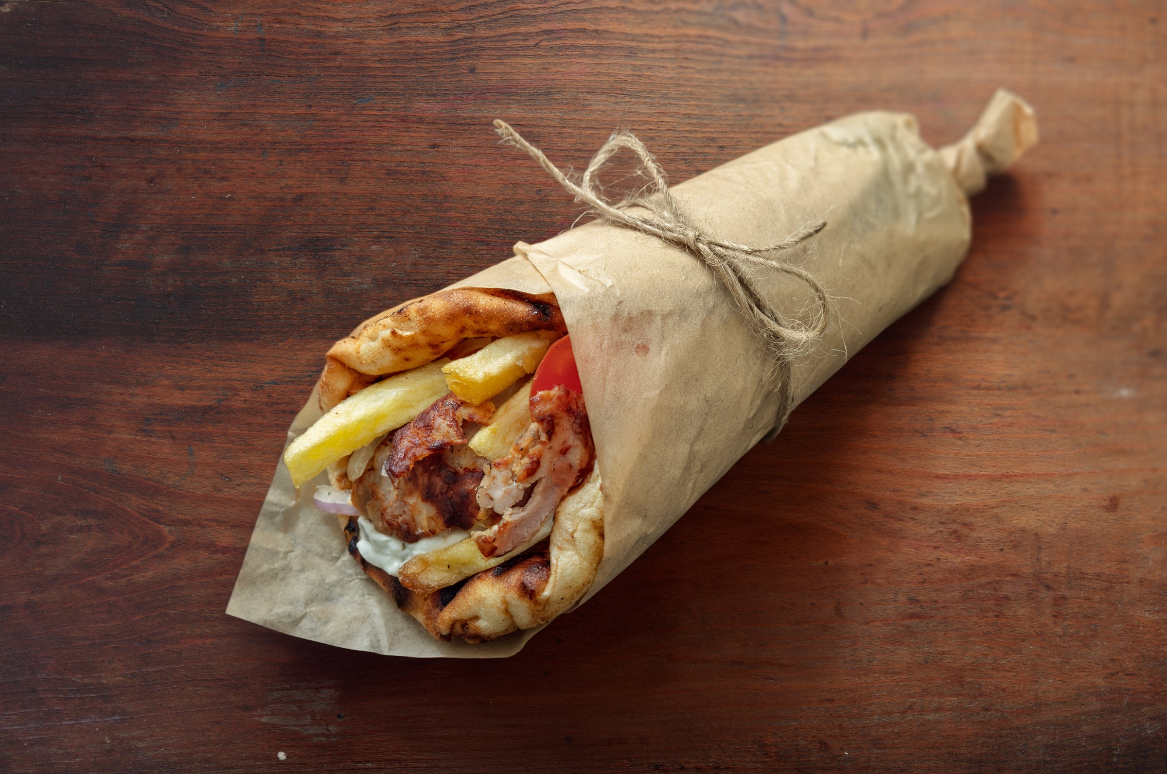 Close-up of a traditional Greek gyros wrap with grilled meat, fries, and vegetables in pita bread, resting on a rustic wooden table under warm natural light, evoking a cozy Mediterranean feel.