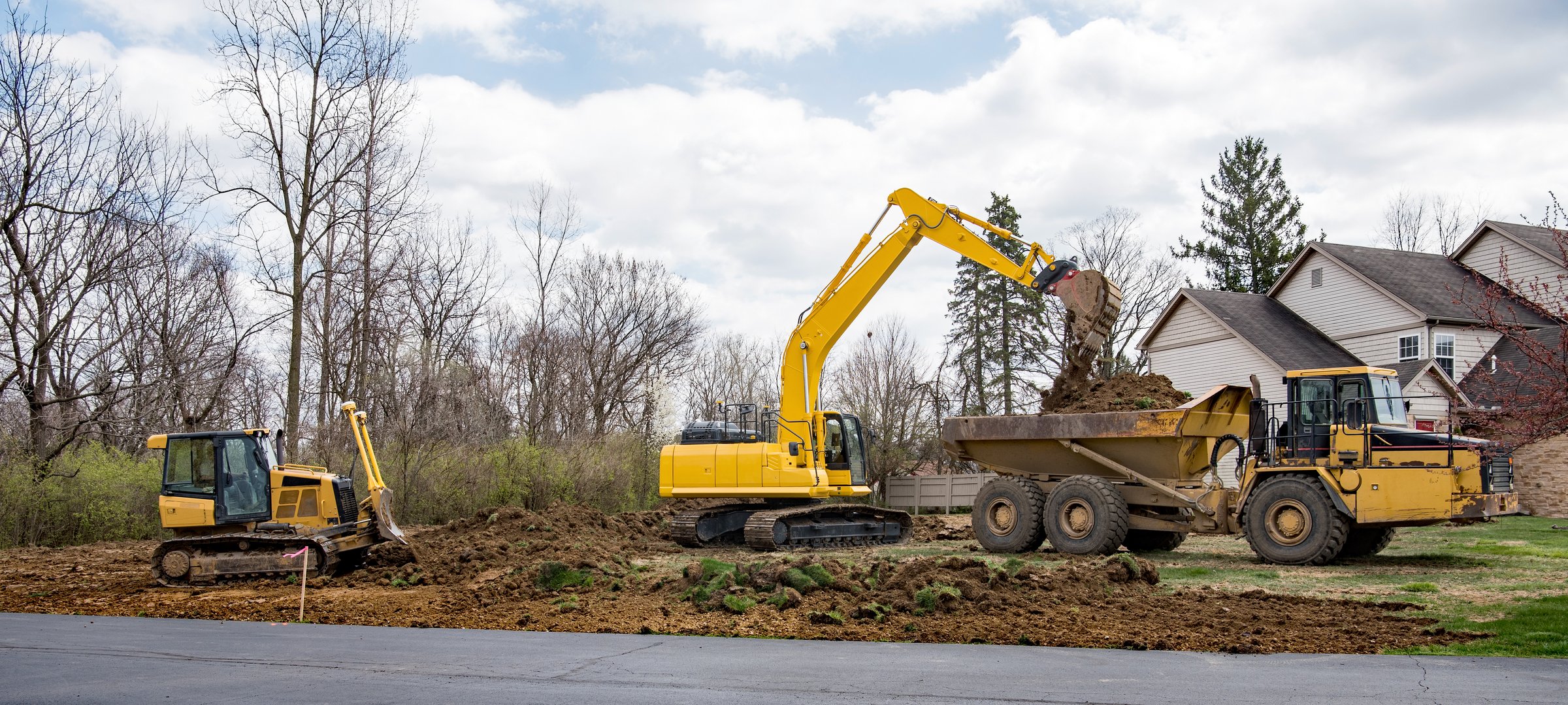 Panorama of bulldozer leveling soil while backhoe is dumping excavated soil into dump truck bed.