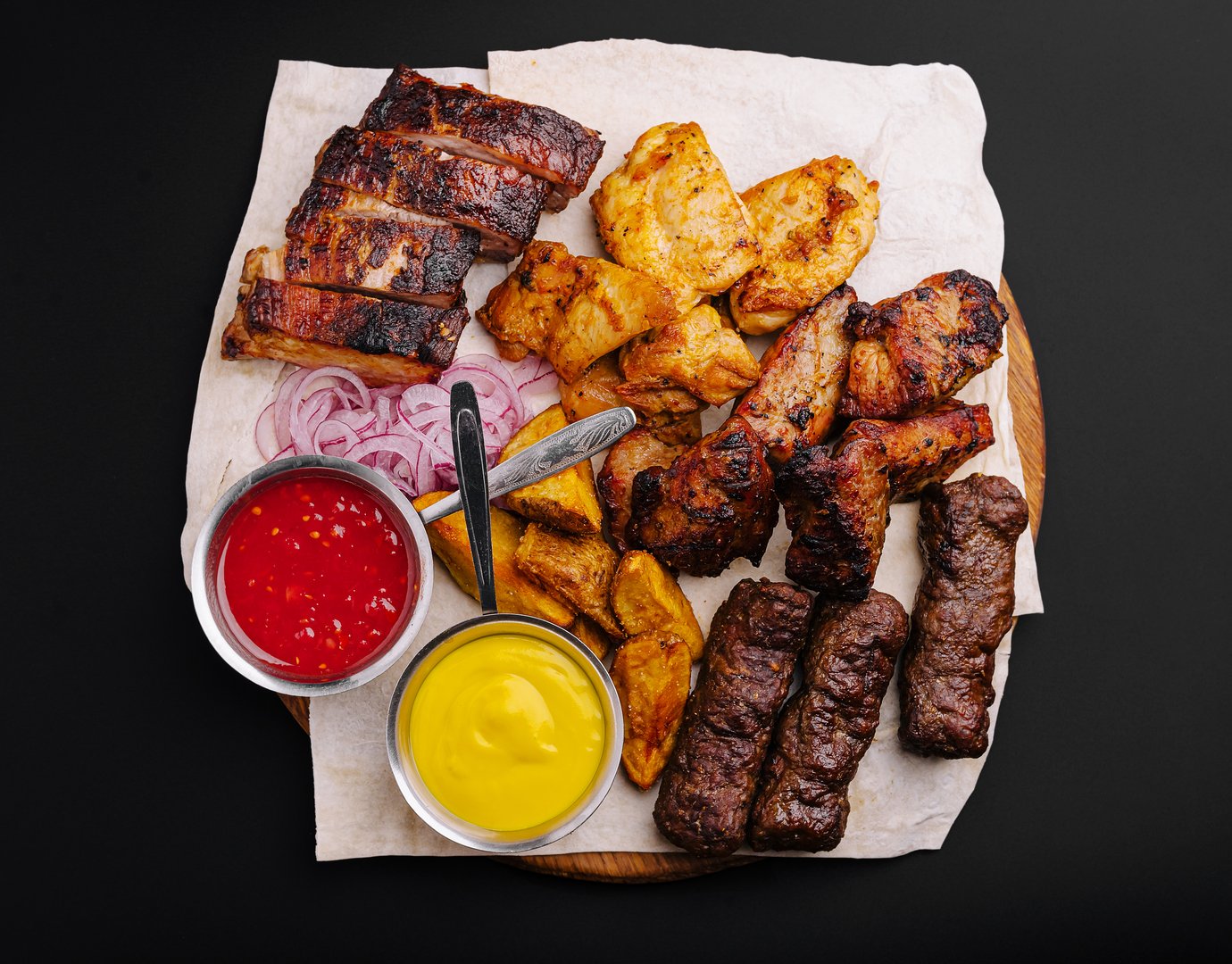 A vibrant assortment of grilled meats including ribs, chicken, and skewers is artistically arranged on a wooden platter. Accompanied by tangy sauces and garnished with onions, it invites sharing.