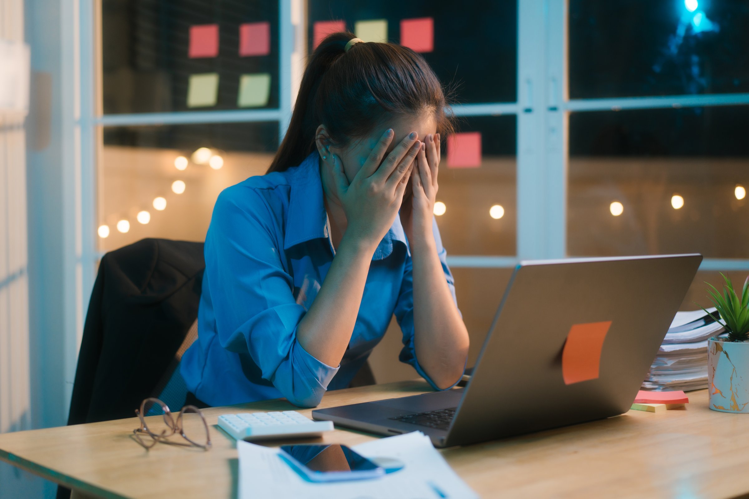 Working late into the night, Asian woman appears stressed and overwhelmed at her desk, rubbing her forehead as if experiencing migraine or headache, surrounded by paperwork and dimly lit office.