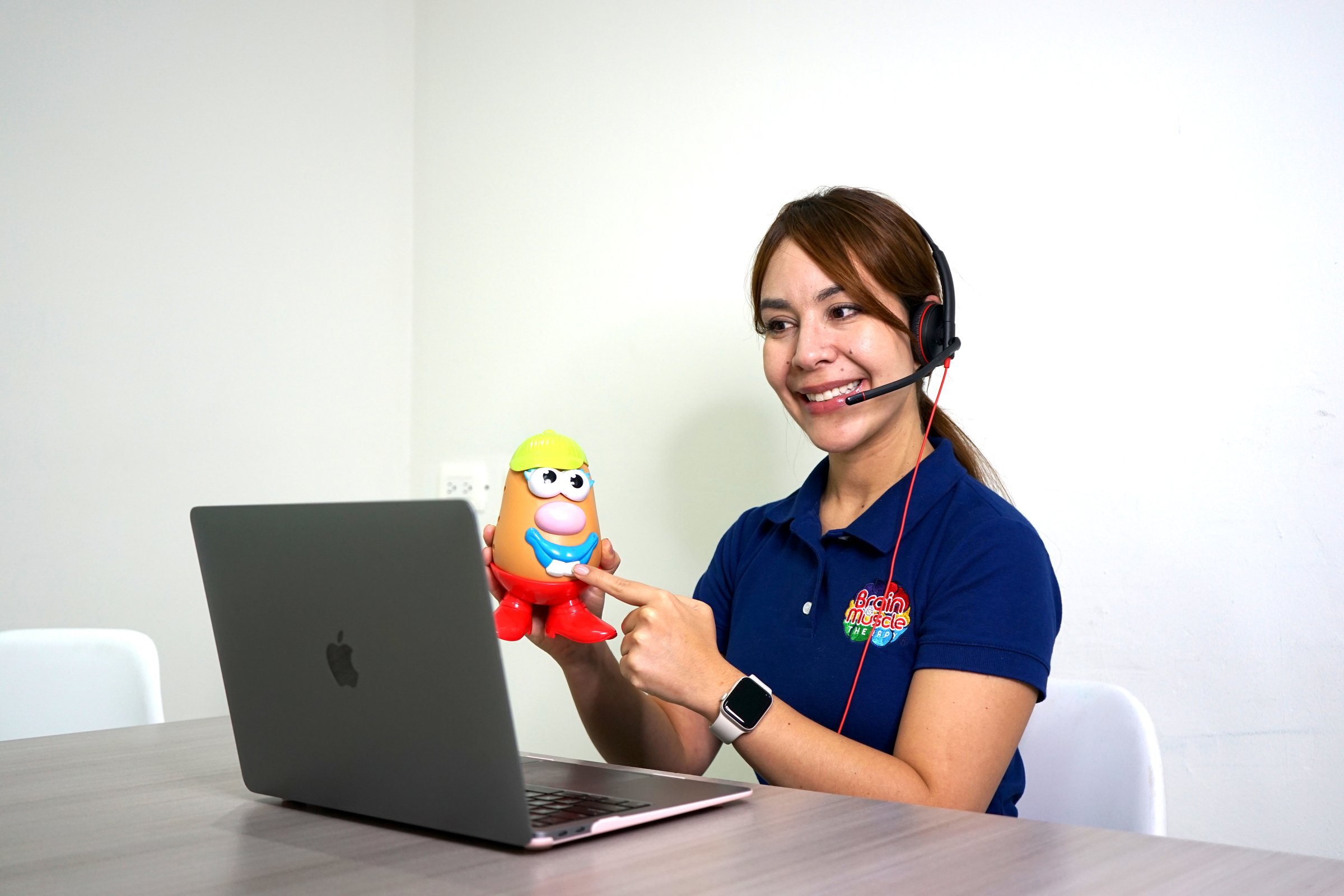 Woman with headset holding a toy, pointing at it, while sitting at a table with a laptop.