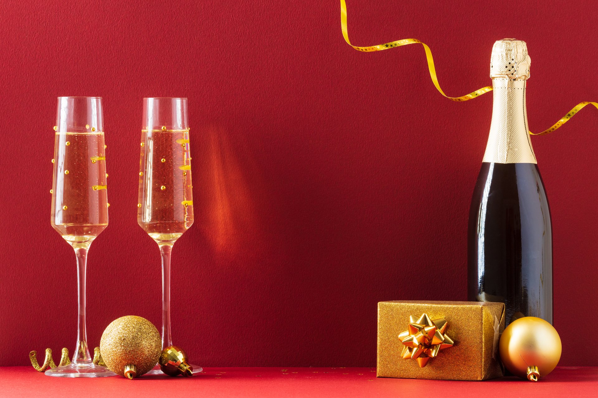 Luxury holiday setup with champagne, gold gift box, and festive ornaments on deep red background. Ideal for New Year, Christmas, and elegant celebration themes.