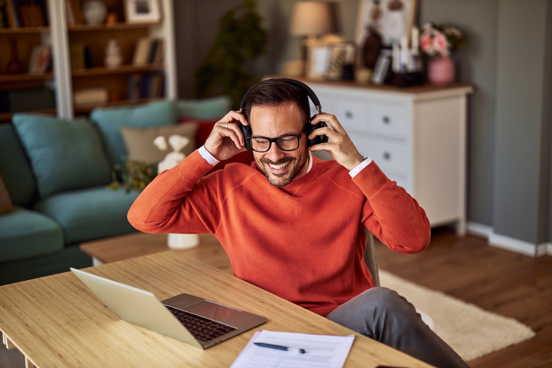 Smiling man in orange sweater working from home, putting on headphones at a tidy desk with laptop and documents.