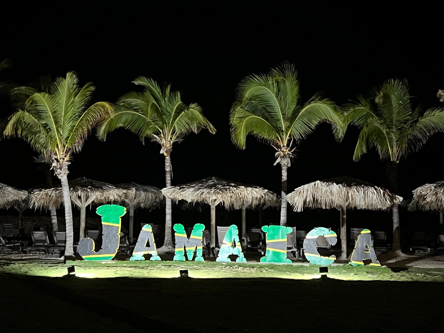Illuminated Jamaica sign with tiki hut umbrellas and palm trees at night.