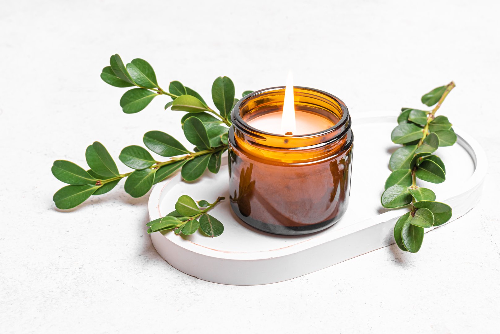 Burning soy wax candle in amber glass jar and green plant leaves on white concrete tray. Natural home decor, spa template and aroma massage concept.