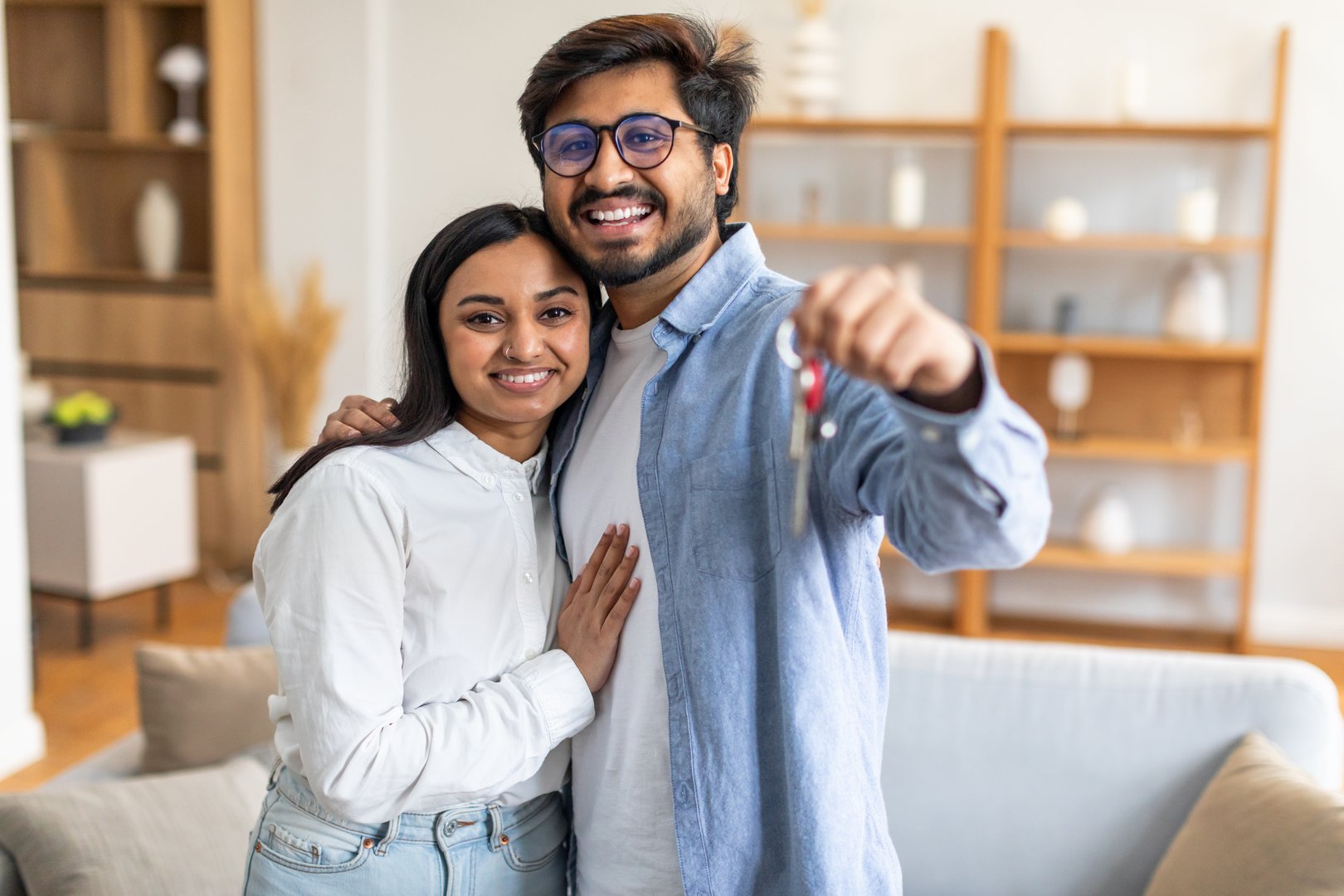 A joyful Indian couple embraces and smiles broadly while holding a set of keys, signifying the purchase of their new home. They stand in a modern, well-lit living room with minimalist decor