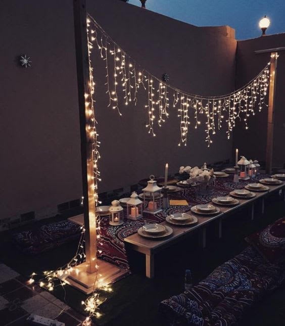 Cozy outdoor dinner setup with low tables, patterned cushions, hanging string lights, and decorative lanterns.