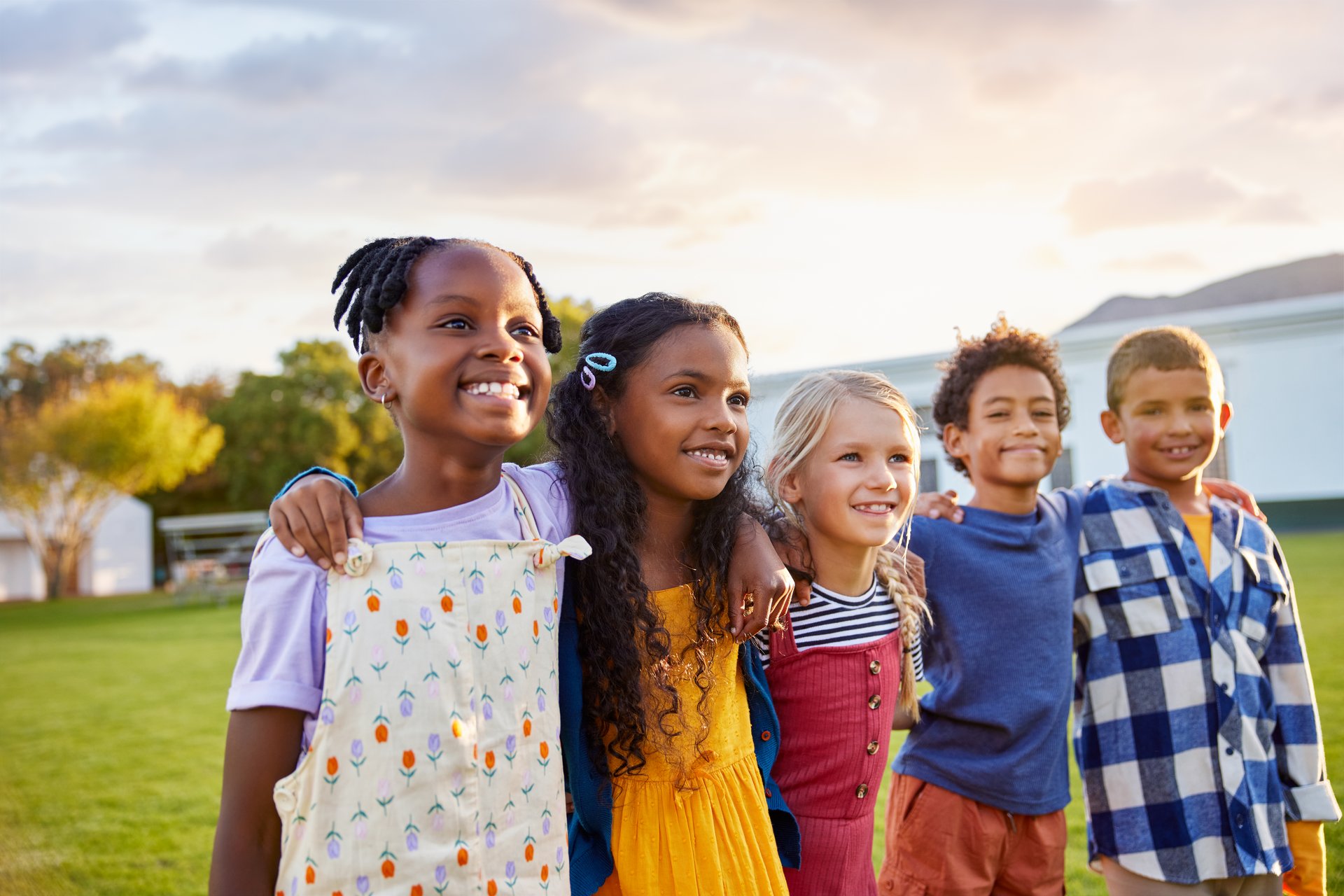 Side view of group of friends standing and hugging while looking away. Small schoolchildren enjoying summer break in outside park. Group of diverse little boys and cute girls with arms around smiling together and think about the future.