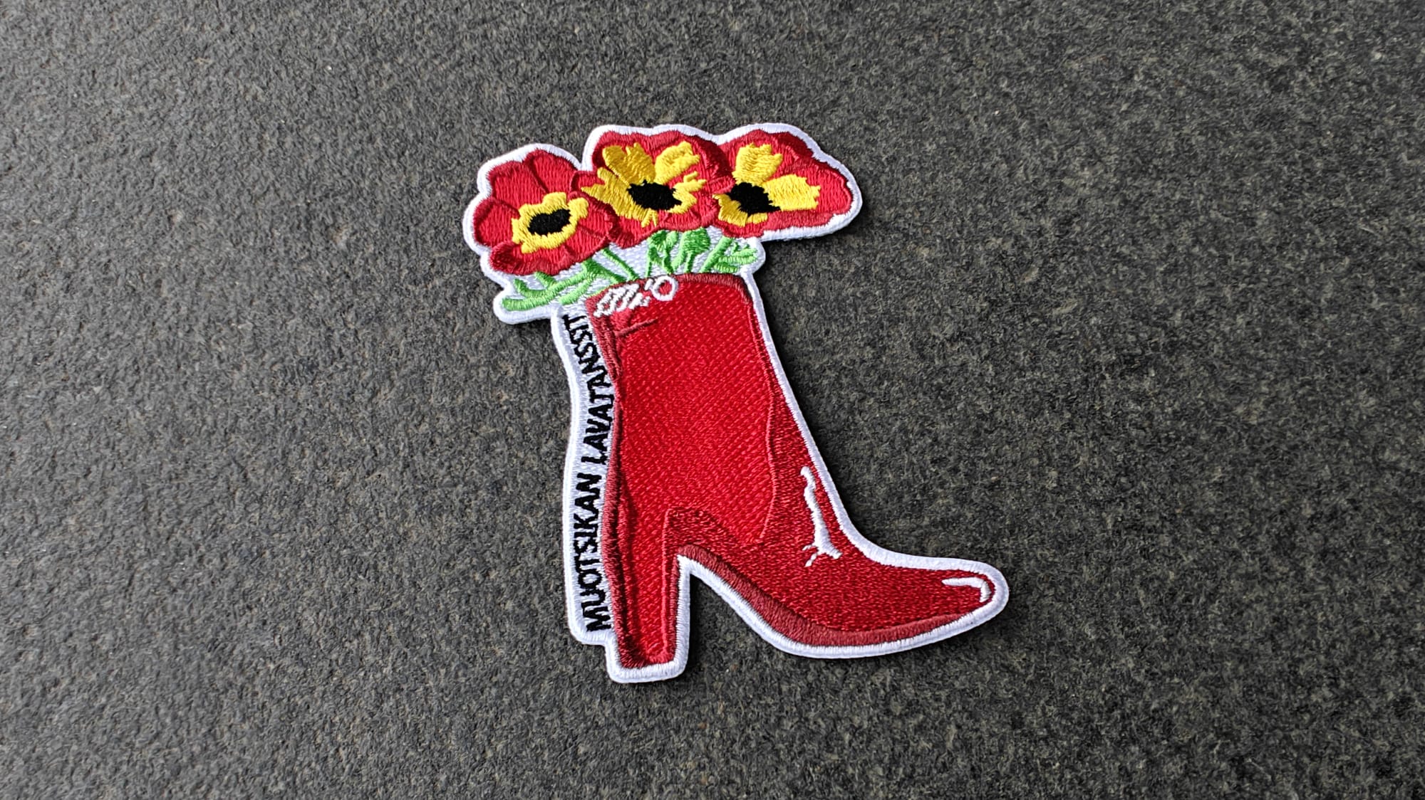 Patch of a red high-heeled boot with red flowers inside, on a gray background. Text MUOTSIKAN LAVATANSSI visible on boot.