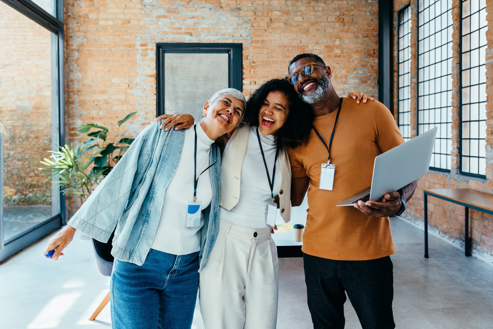 A group of diverse colleagues share a joyful moment in a modern office with brick walls and abundant natural light, representing teamwork and camaraderie.