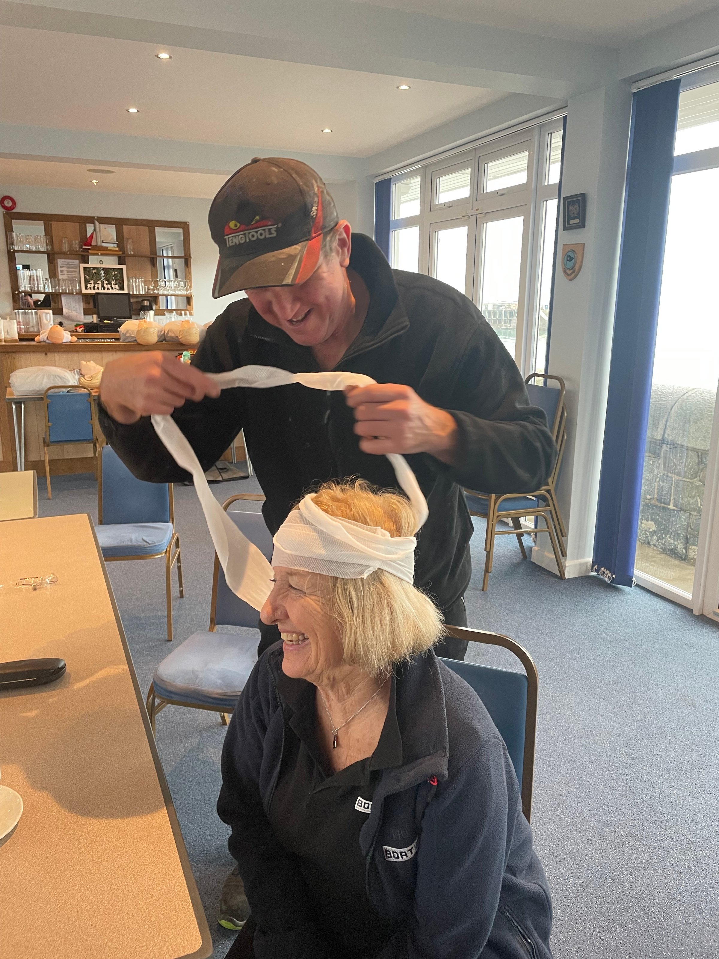A smiling woman sitting while a man playfully wraps a bandage around her head in a casual indoor setting.
