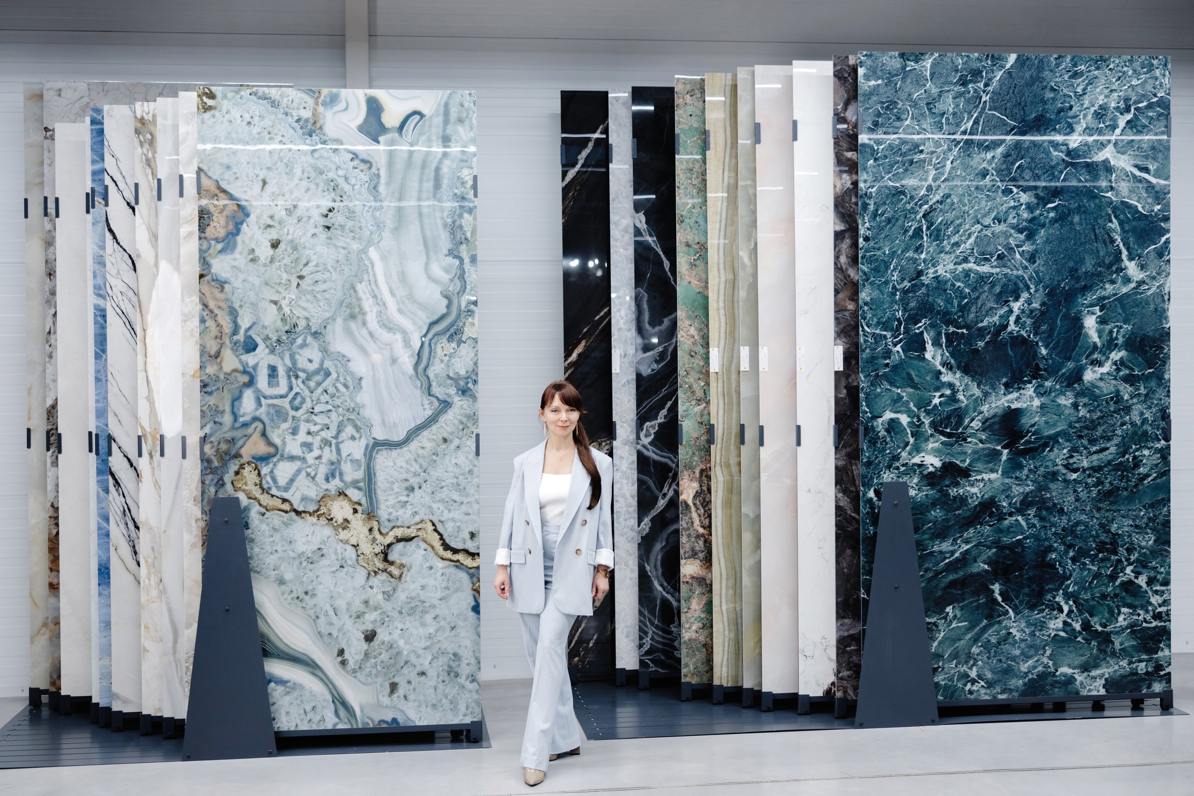 A woman in a light blue suit stands in front of a display of marble tiles. The tiles are arranged in rows and columns, showing a variety of colors and patterns.