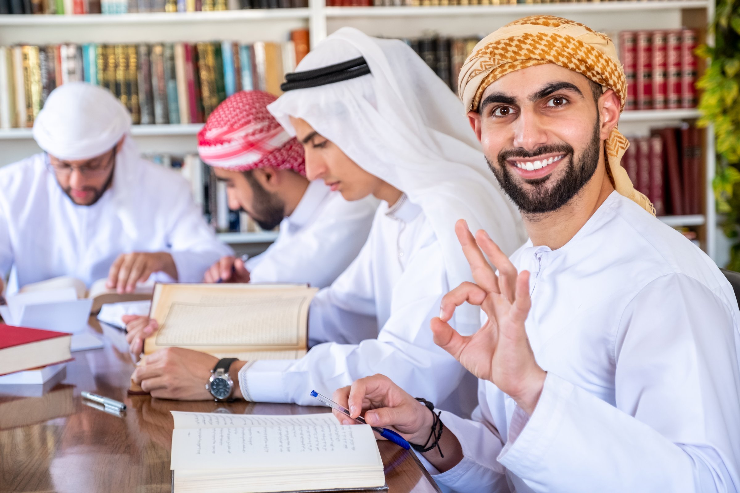 A group of Middle Eastern students wearing traditional attire study together, surrounded by books in a library. One student smiles and gestures approval. The atmosphere is collaborative and focused
