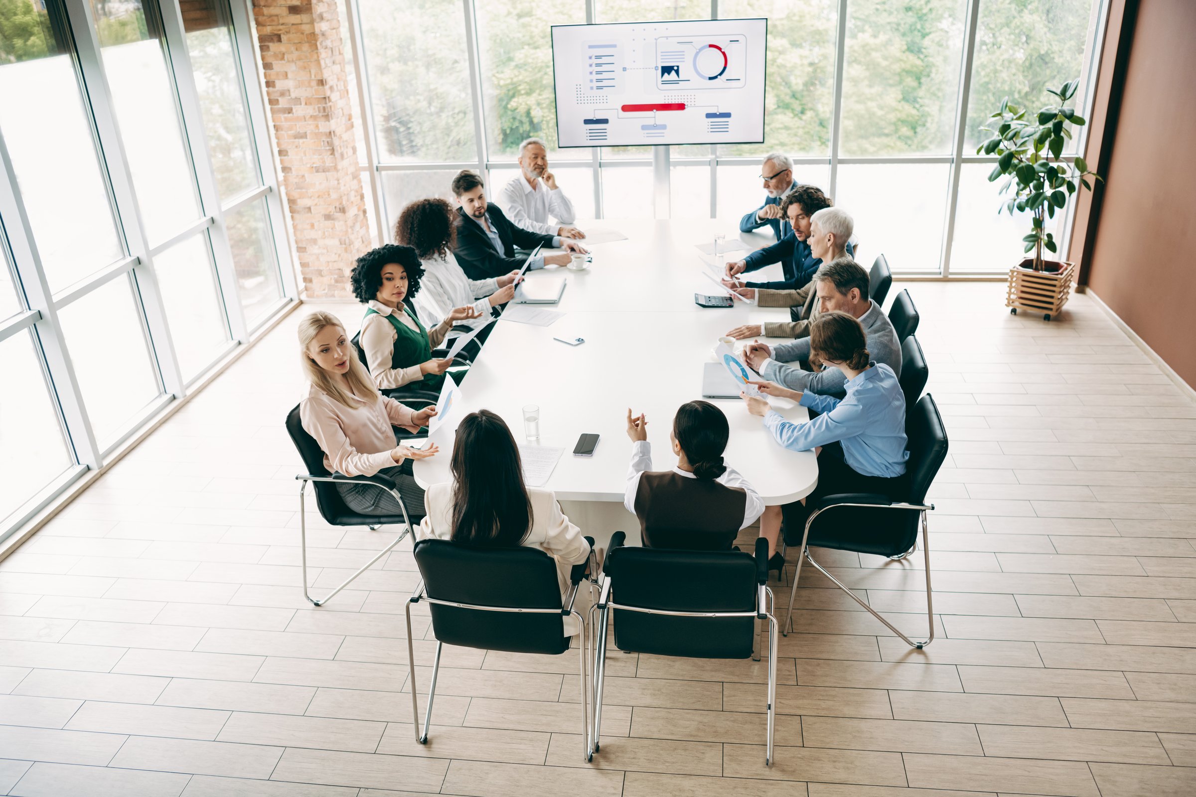 A professional team of colleagues participating in a corporate meeting in an office setting. Discussing strategies and plans around a table with a presentation screen displayed.