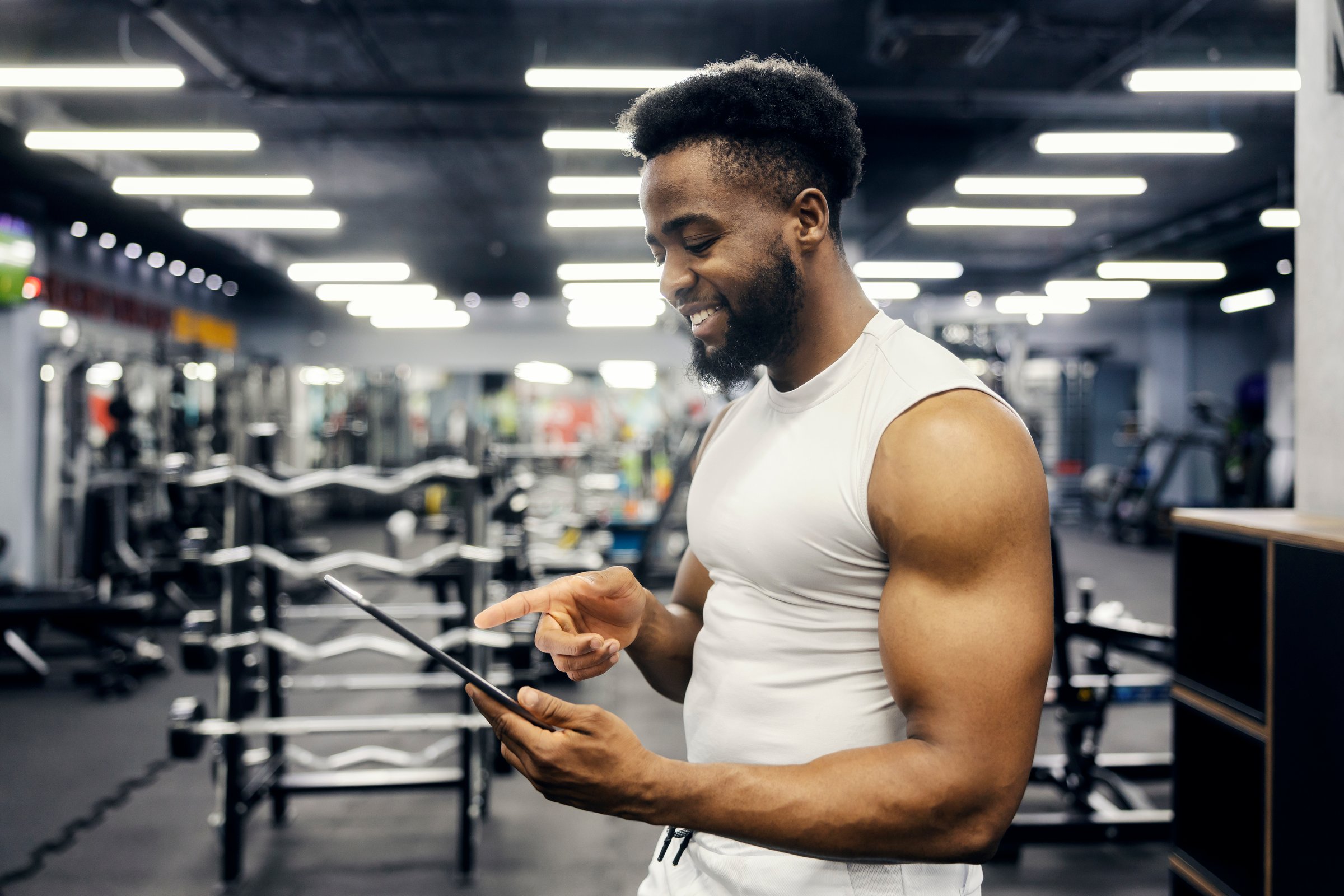 Young adult man smiling while using a digital tablet for fitness tracking and workout planning in a modern gym