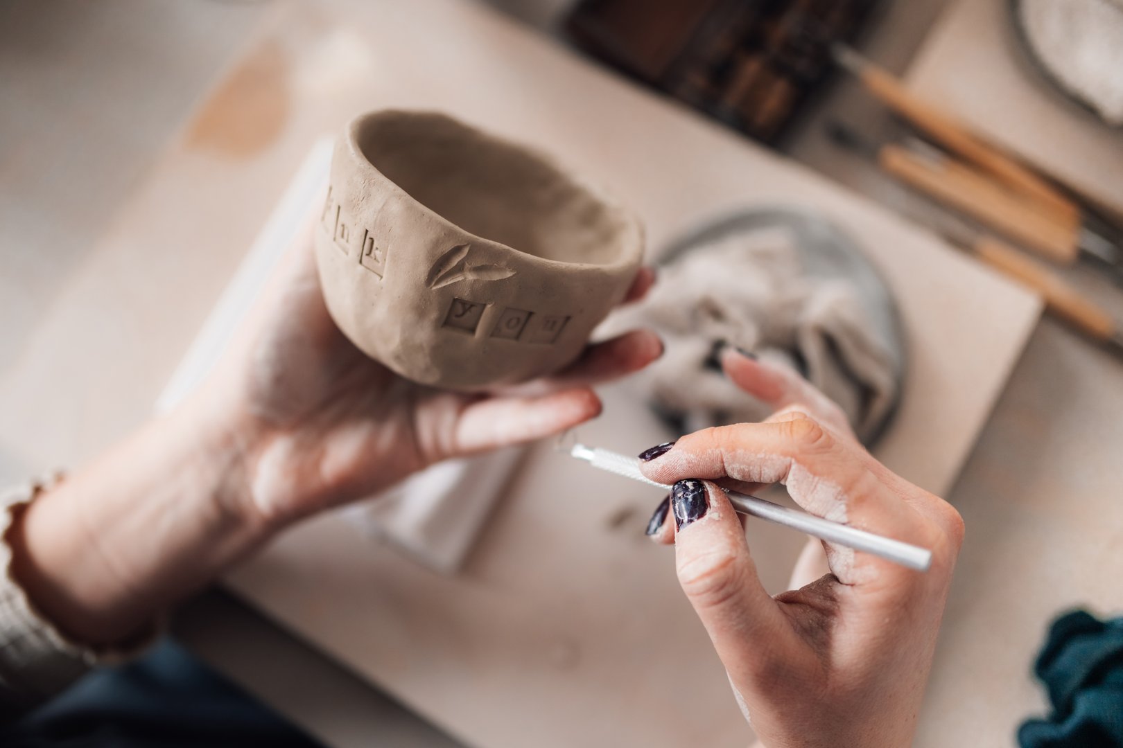 Close up of craftswoman's hands using pottery tool for decorating earthenware at pottery workshop. Cropped picture of unrecognizable creative pottery artisan using decorative sharp tool on clay mug.
