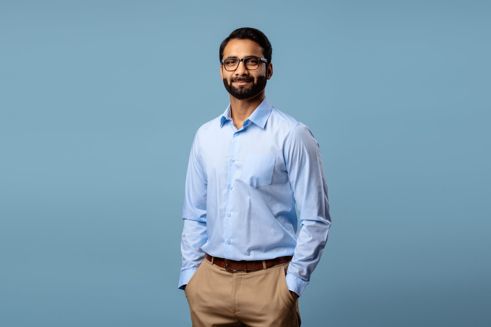 Portrait of confident Indian businessman wearing eyeglasses, hands in pockets, looking at camera, standing isolated on blue background. Advertisement concept
