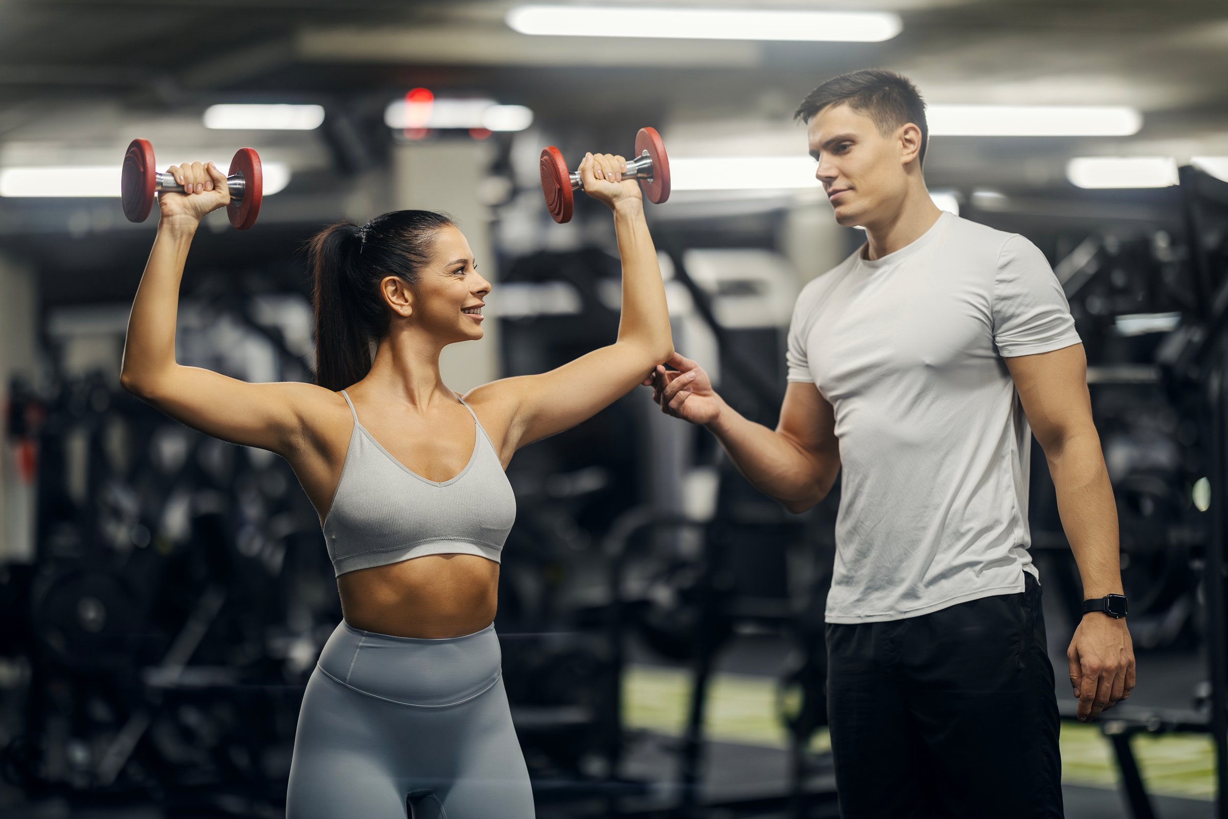 Woman lifting dumbbells with a male personal trainer helping her in a modern gym, focusing on strength and fitness