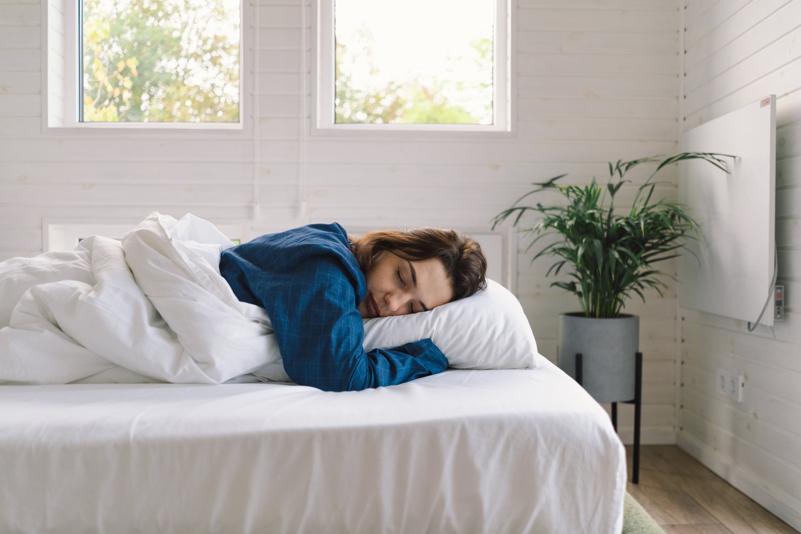 A woman lies comfortably on her back in bed, wearing a blue shirt, smiling as she stretches. Sunlight gently illuminates her peaceful expression, enhancing a serene morning atmosphere.