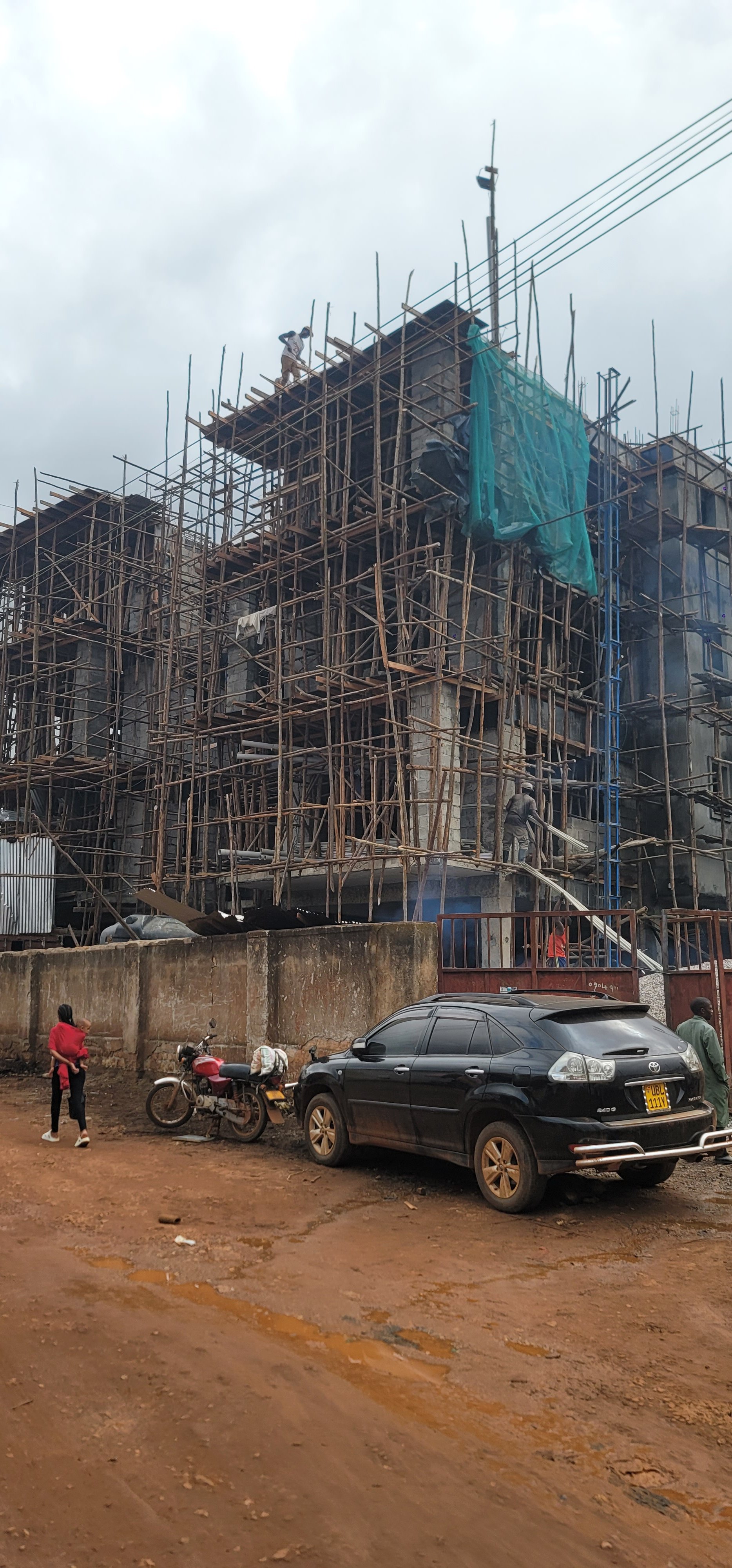 A multi-story building under construction with scaffolding, cars parked nearby, and a few people on the street.