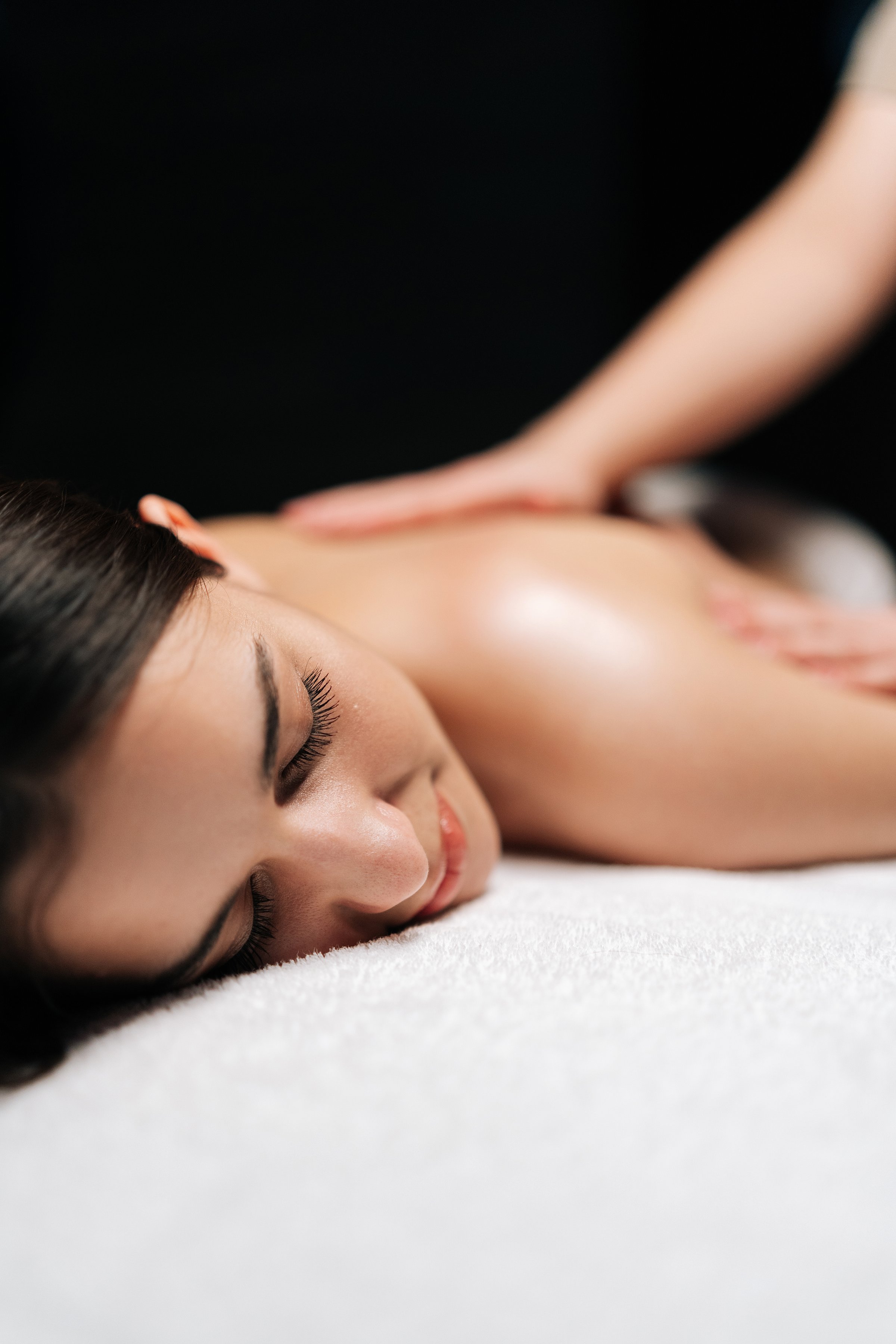 Closeup vertical shot of carefree woman client relaxing during back massage in spa salon with dark interior. Professional massage therapist providing therapeutic back massage to relaxed female.