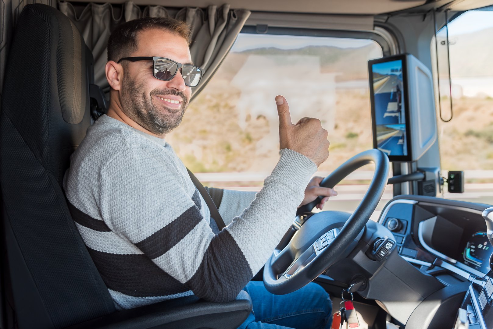 Young truck driver with a happy face and a thumbs-up gesture clearly signaling OK while driving a heavy vehicle.