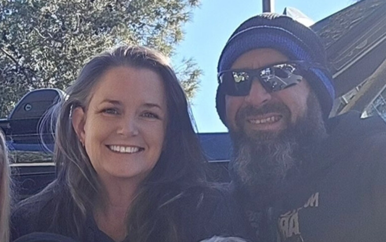A smiling woman and a bearded man wearing sunglasses and a beanie pose outdoors, with trees and a vehicle in the background.