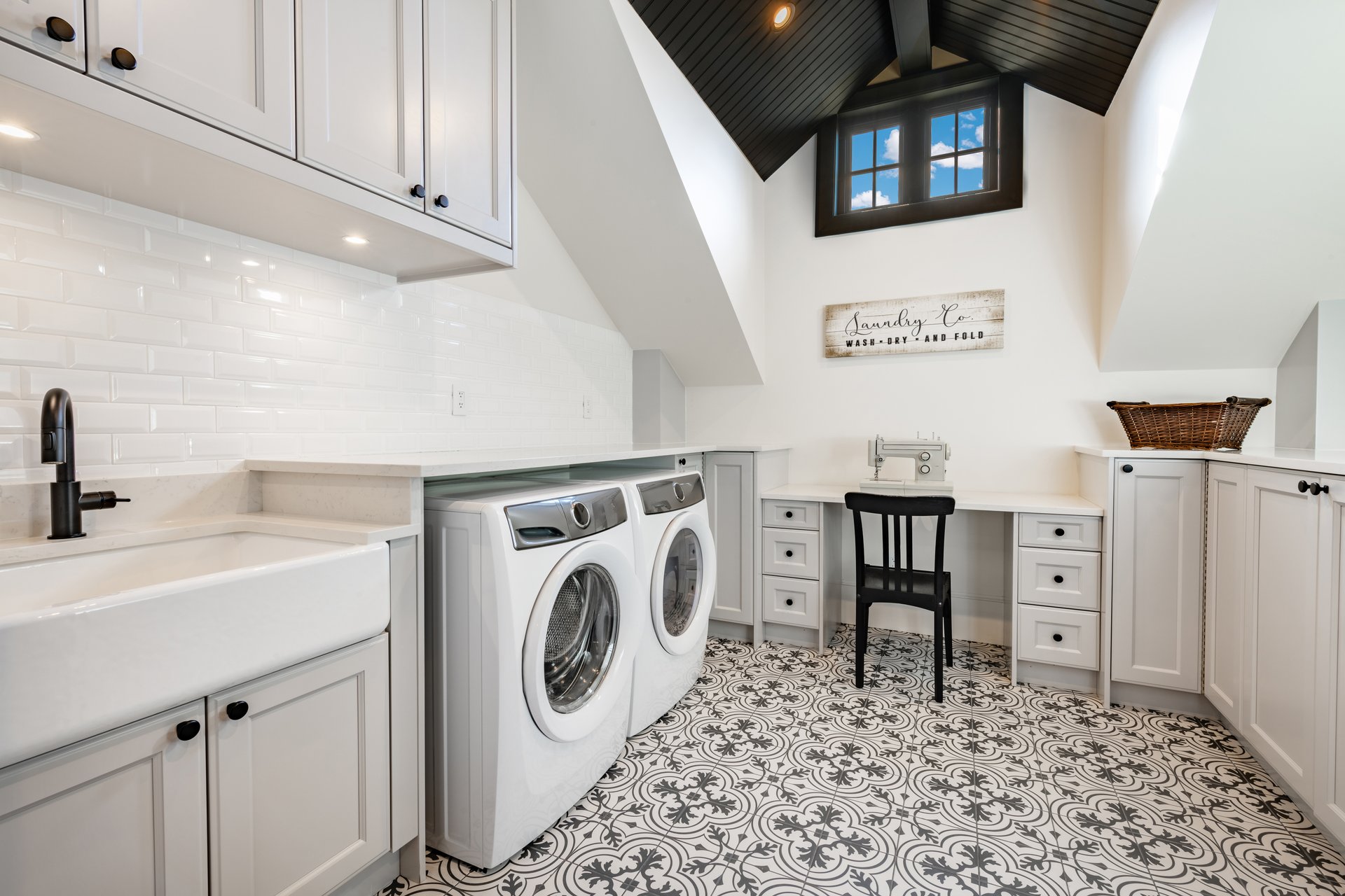 Laundry room interior with patterned tiled linoleum floor farmhouse sink and desk with sewing machine washer and dryer vaulted ceiling with wooden beams white cabinets and countertops spacious and luxurious domestic housekeeping space