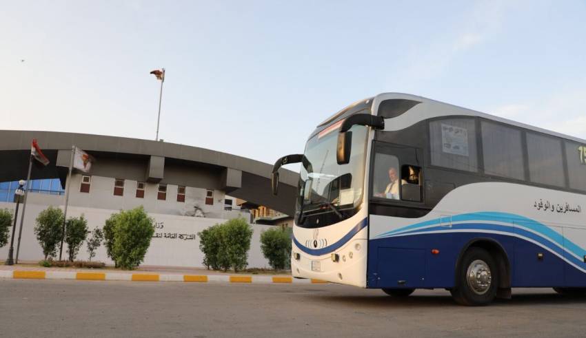 A blue and white bus parked near a building with Arabic text and flags, under a clear sky.
