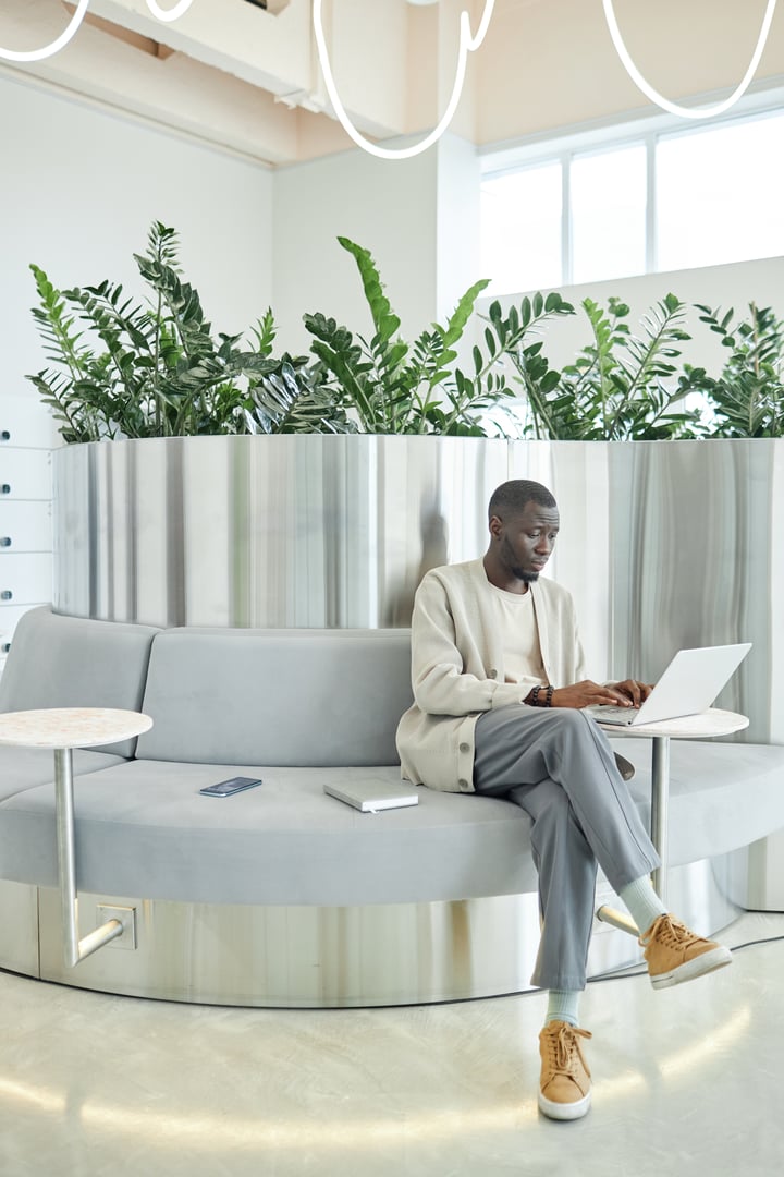 Vertical shot of African American businessman typing on laptop in lounge zone while working on designer furniture in coworking space featuring biophilic interior design