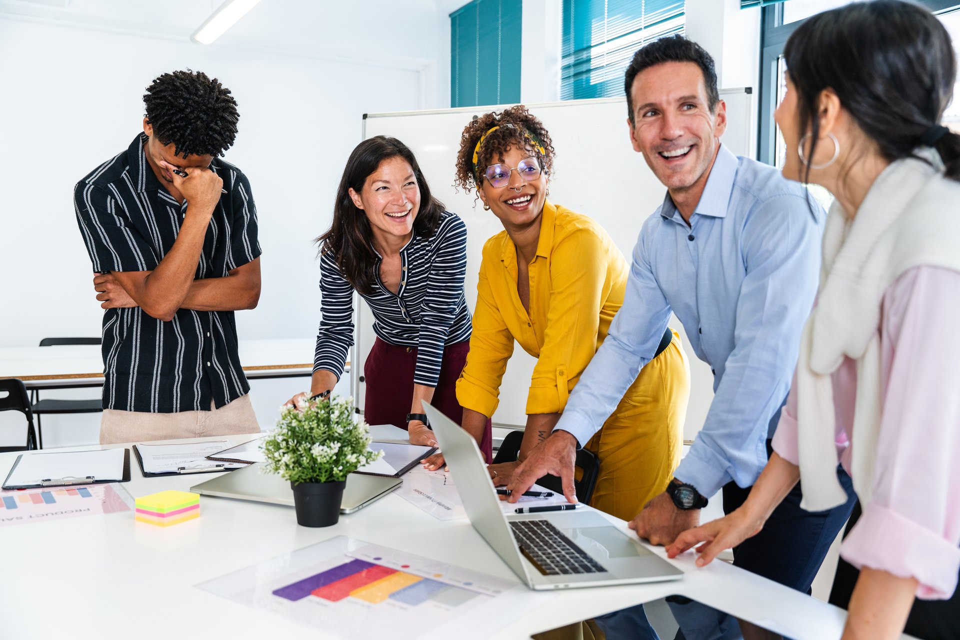Multi-ethnic business team collaborating on a project, sharing ideas and laughing together around a table with laptop and documents in a modern office