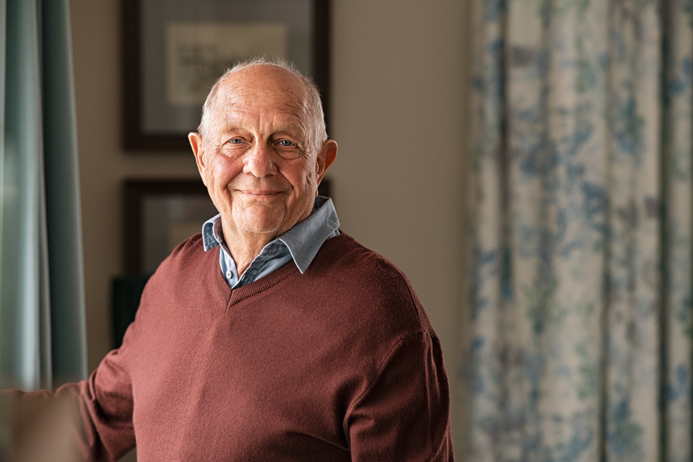 Portrait of happy retired senior man standing at home near window. Satisfied old man in casual clothes looking at camera and smiling while standing near the window.