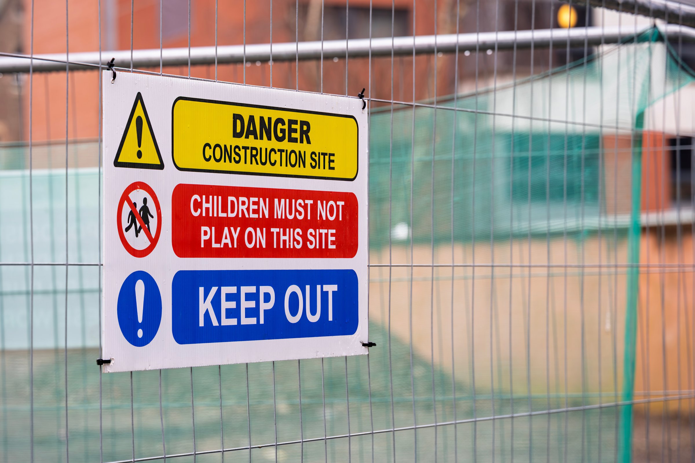 Blue, Red, Yellow and White Children Keep Out Construction Site Warning Sign on Temporary Heras Fence Panel with Green Netting and Blurred Background