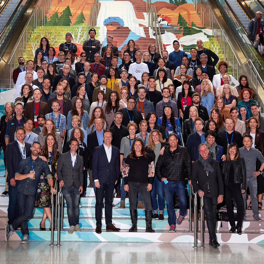 A large group of people, both sitting and standing, pose on stairs decorated with a landscape mural in an indoor setting.