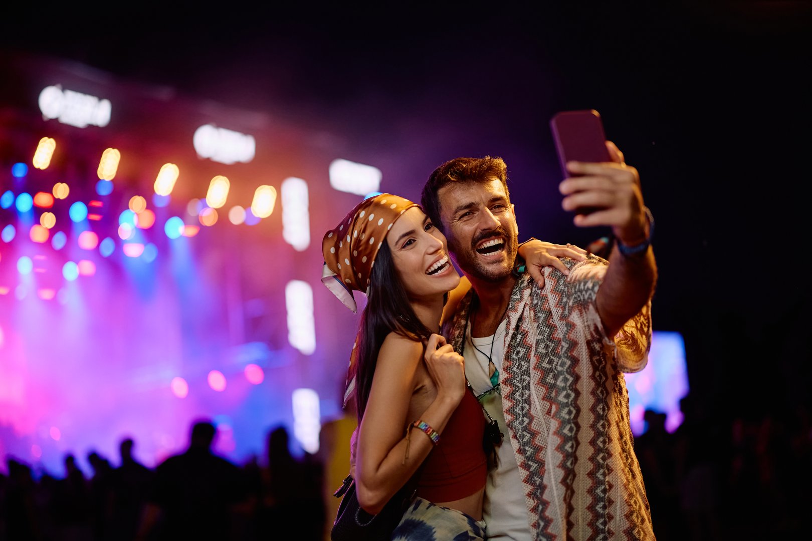 Cheerful couple using cell phone while taking selfie on summer music festival at night. Copy space.