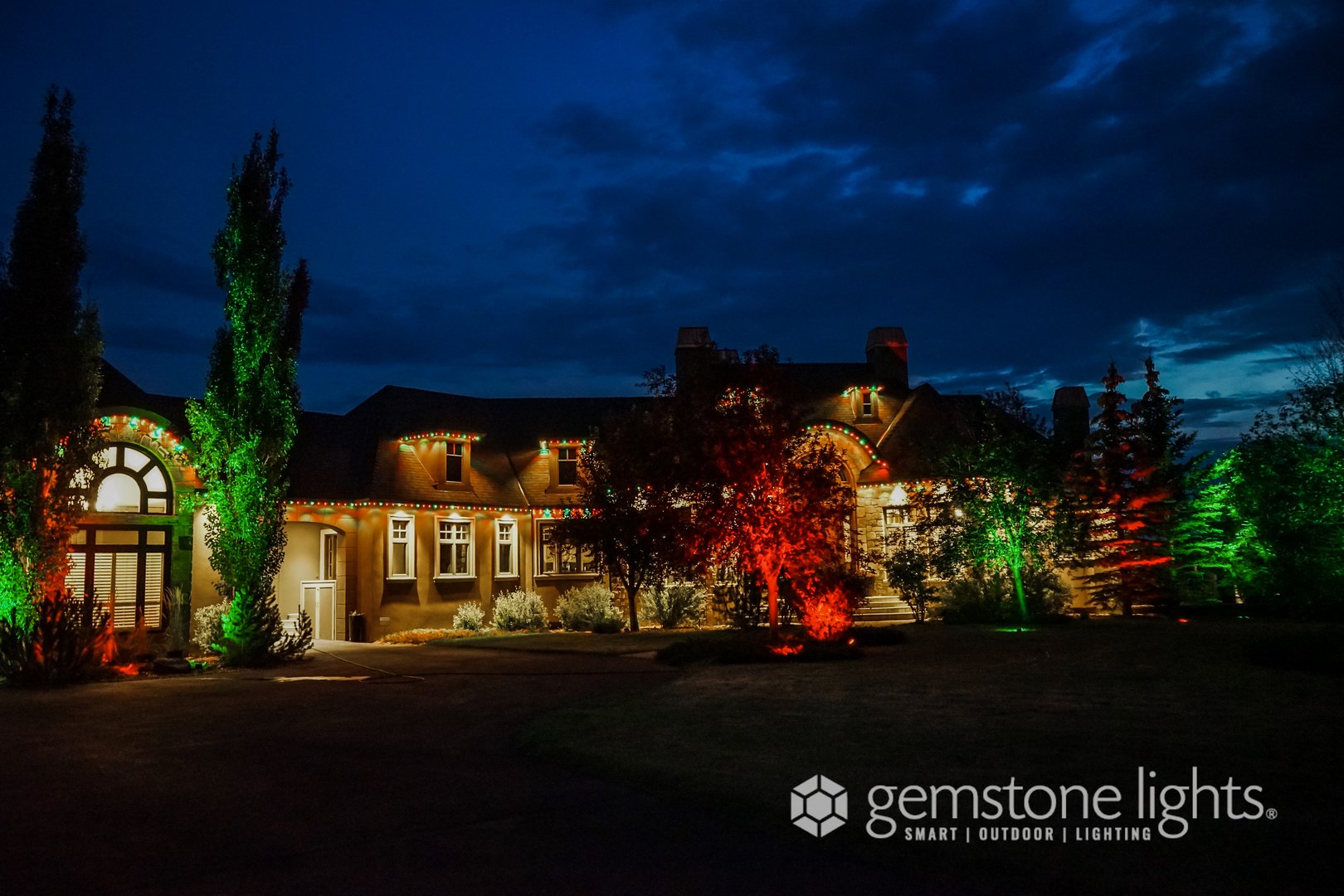 Large house illuminated with colorful lights at night, with trees and sky in the background. Gemstone Lights logo in corner.