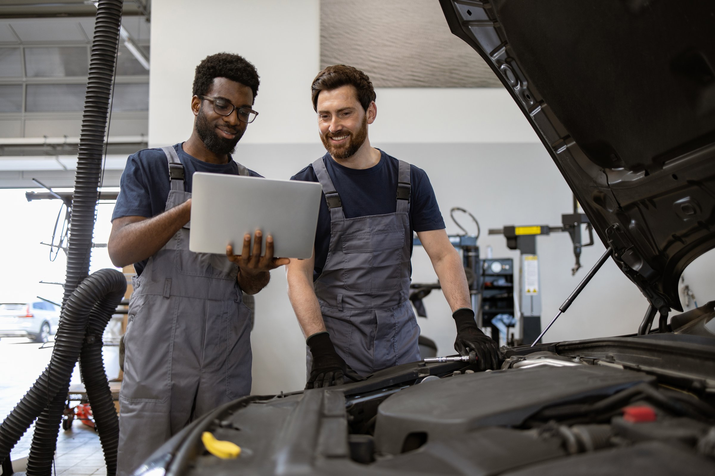 Two mechanics working together to diagnose car issue using laptop in repair garage. Teamwork and technology in automotive service.