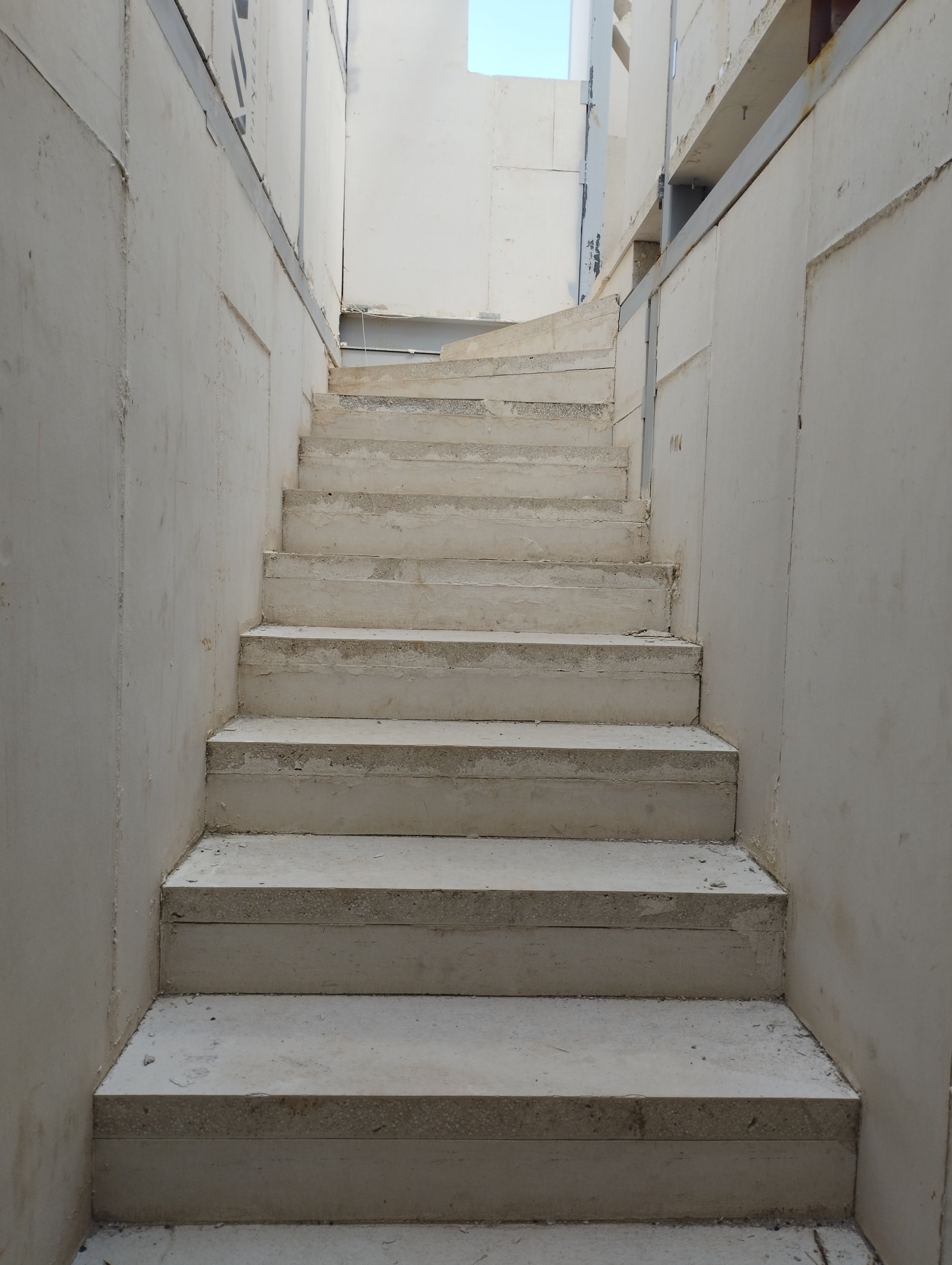 Concrete staircase with white walls on both sides, leading upwards with a patch of sky visible at the top.