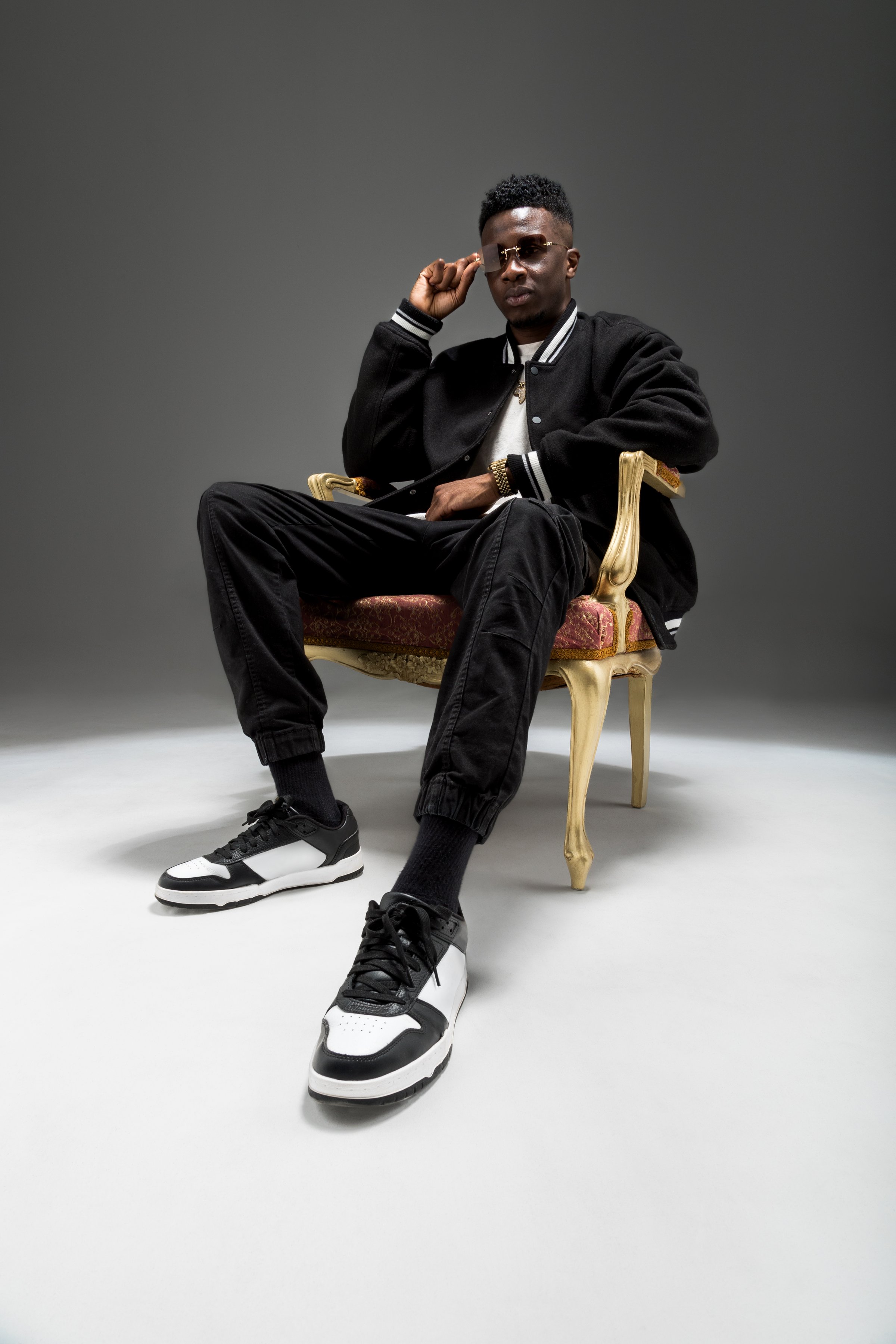 A stylish man sits confidently on an ornate chair, dressed in modern attire against a neutral backdrop.