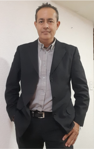 Man in a black suit with a gray shirt standing against a white wall, hands in pockets, with a slight smile.