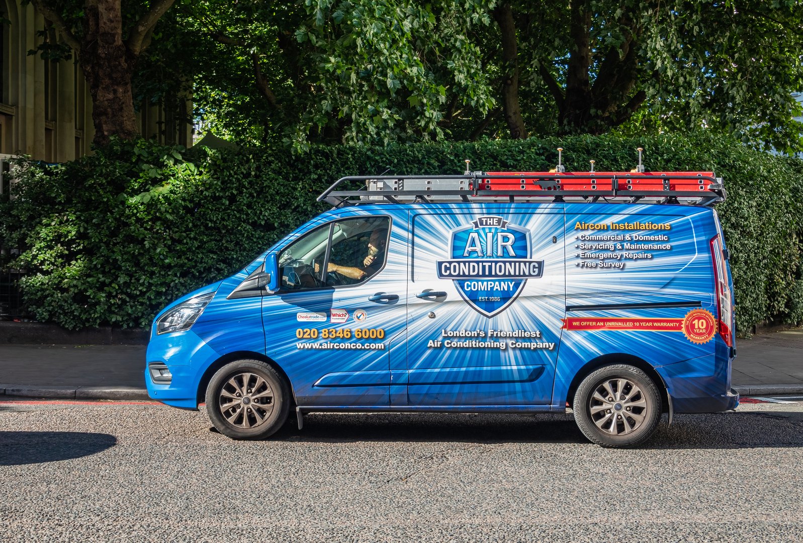 London, UK - July 5, 2022: Closeup, The Air Conditioning Company Van in Albany street, White and yellow on blue, backed by green foliage. Private company. Red ladder on roof.