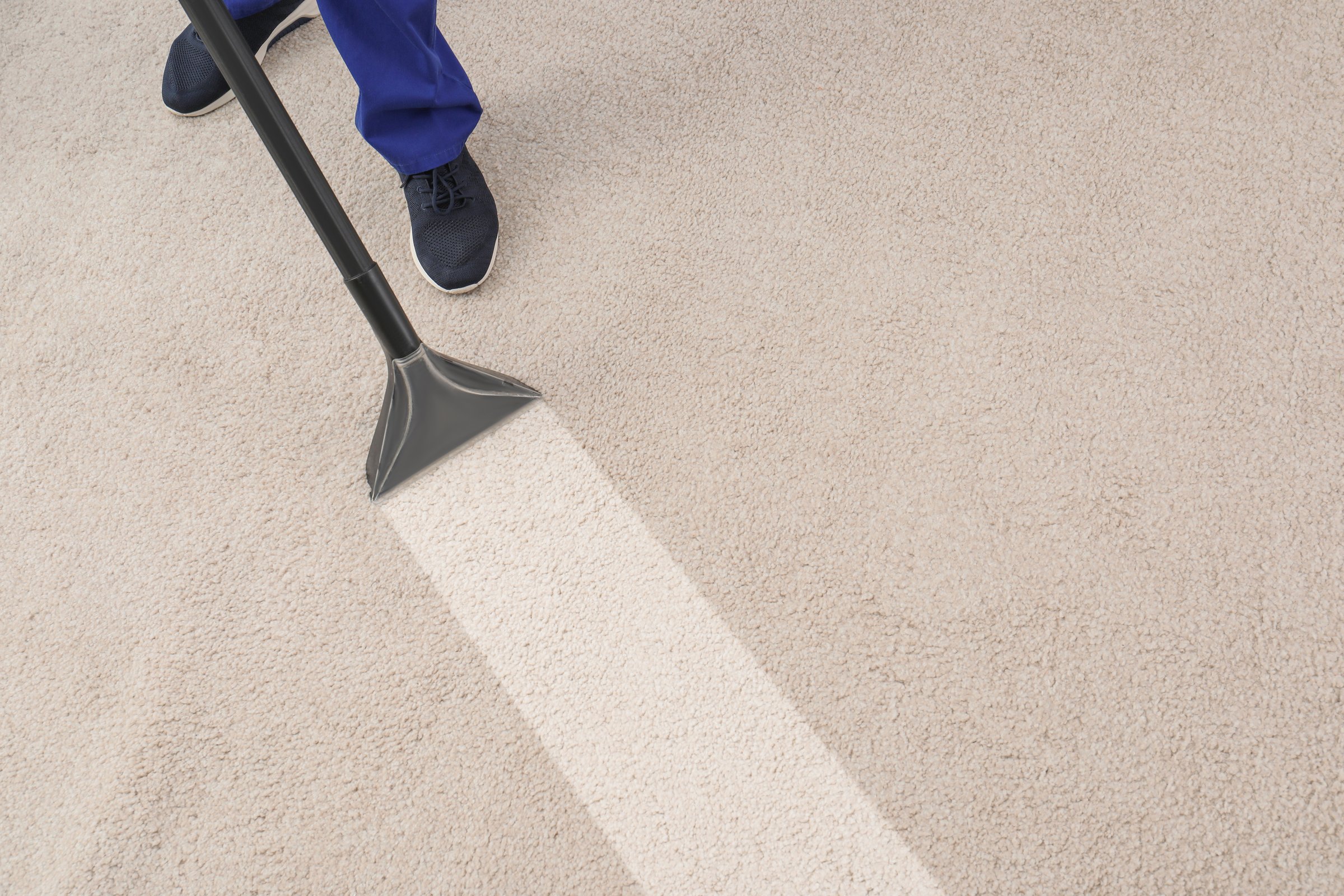 Overhead view of a professional cleaner operating an industrial extractor tool on a beige carpet. The striking contrast between the clean, wet strip and the dirty area highlights professional deep cleaning services, maintenance, and improved home or commercial hygiene.