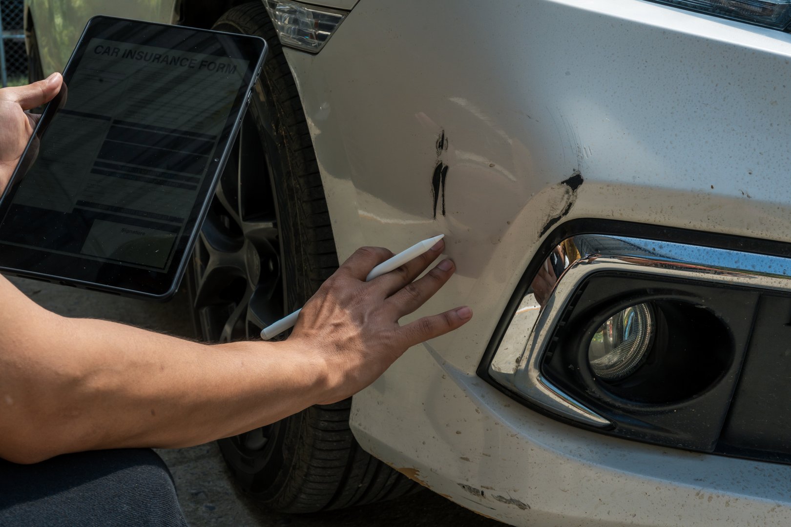 A businessman stands next to his damaged vehicle after an accident, holding a clipboard and examining the scene. An insurance agent arrives to assess the damage and start the claim process.