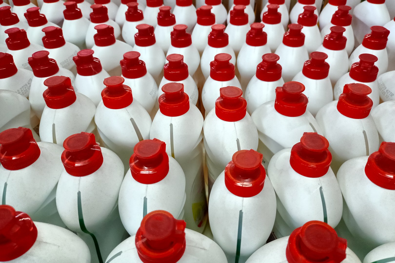 Close-up view of multiple white plastic bottles with red caps, arranged in neat rows, likely containing liquid detergent or cleaning products.