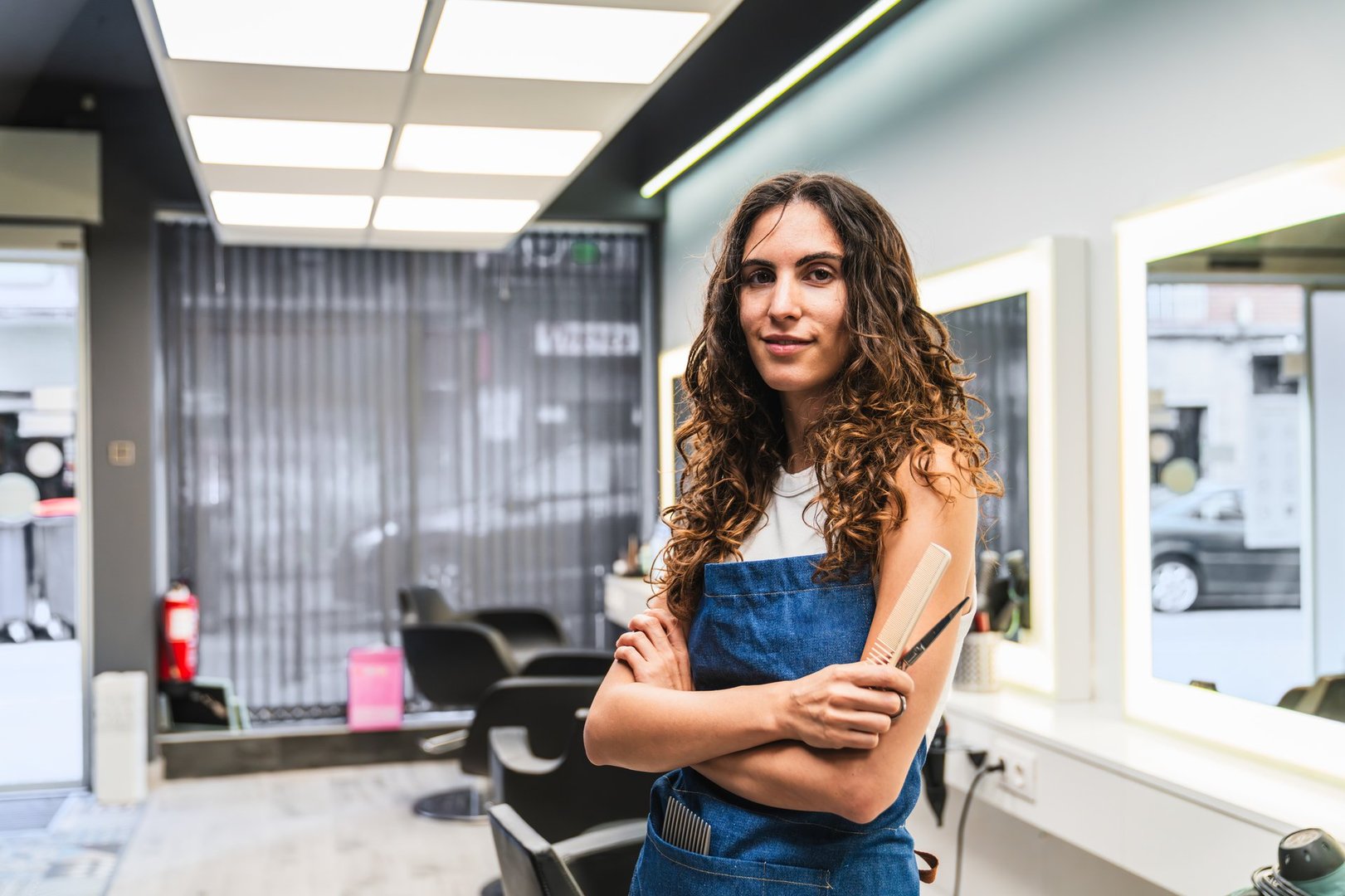 Hairdresser woman wearing an apron, holding scissors and a comb, standing inside her modern hairdressing salon