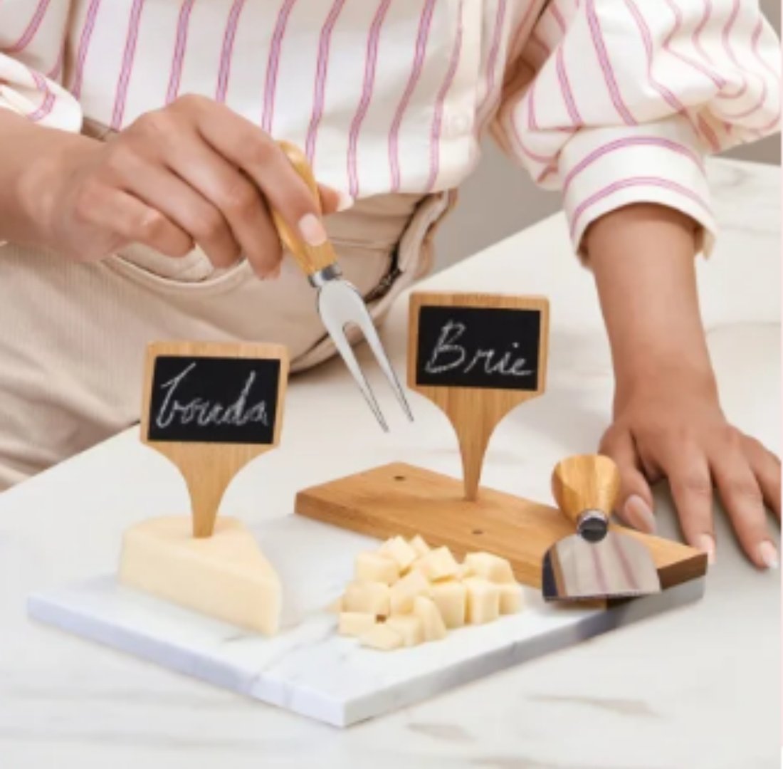 Person arranging cheese with signs on a marble board, using a fork and knife, labeled Toredo and Brie.