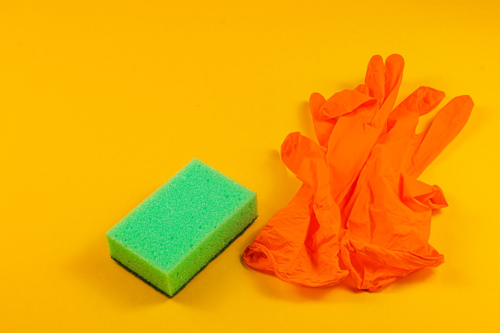 Bright yellow background features a green cleaning sponge alongside orange rubber gloves, creating a vibrant and cheerful atmosphere for cleaning tasks.